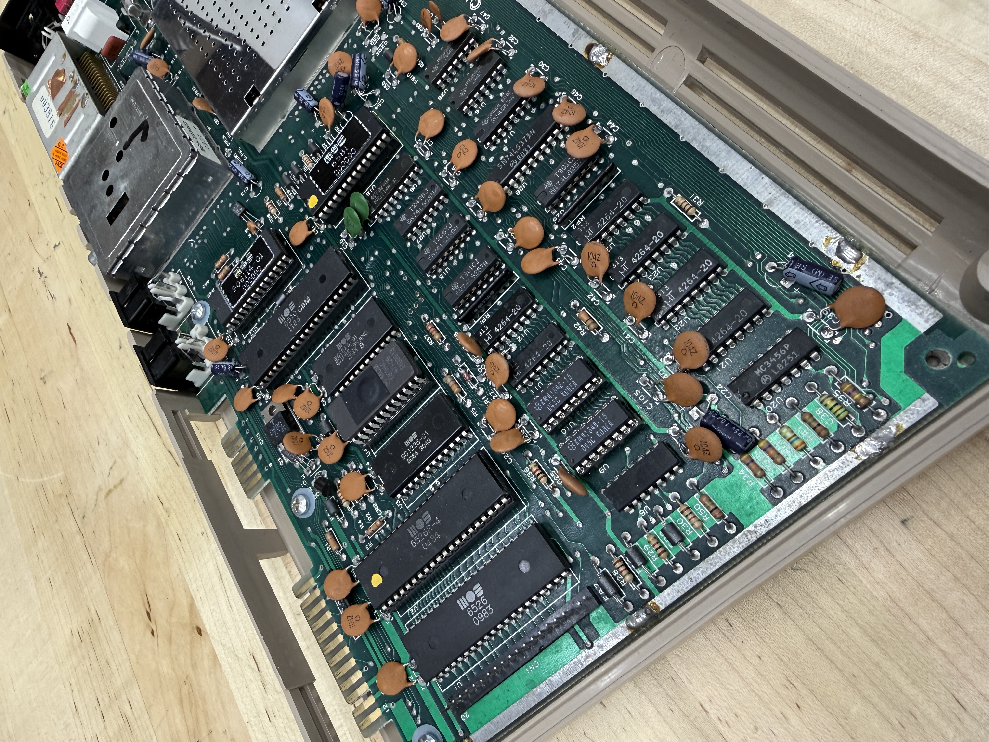 A three-quarters view of a 250407 C64 motherboard in its bottom case shell, top shielding removed, showing the orange disc capacitors, DIP chips, and edge connectors