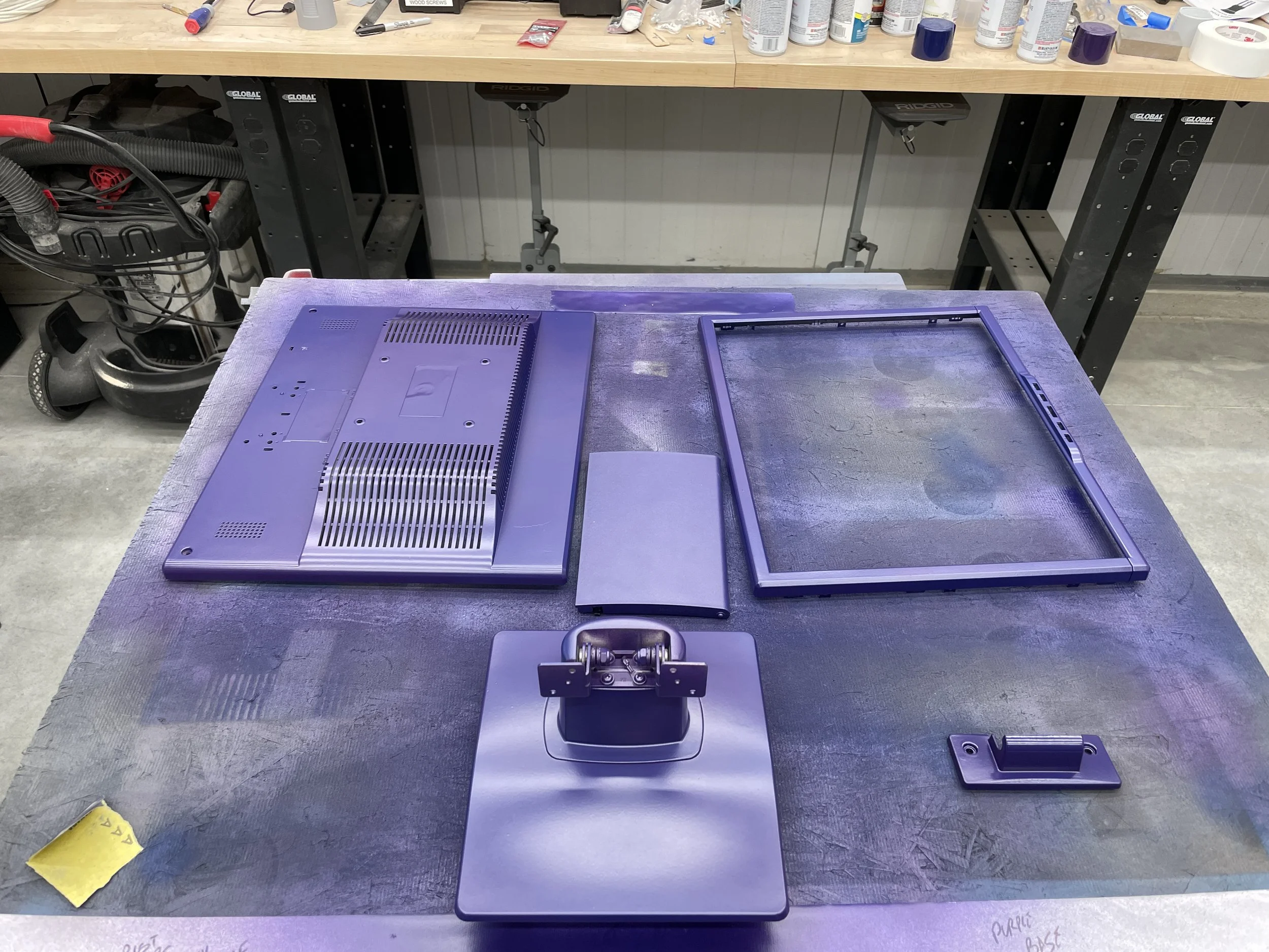 The disassembled monitor parts — rear chassis, bezel frame, stand, and side bracket — painted in Indigo Purple/Blue and curing on the workbench